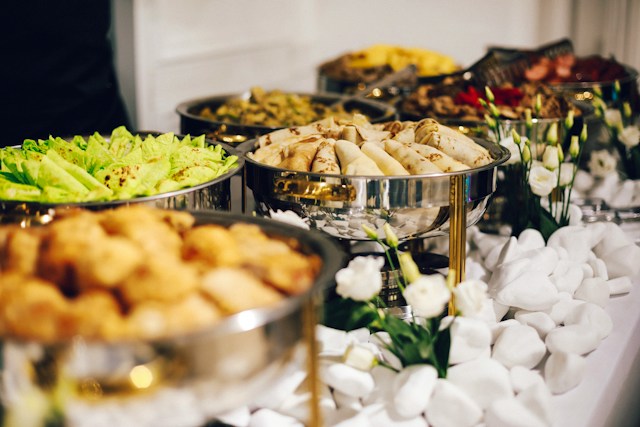 A tasting table laid with Rajasthani dishes in brass and copper serveware on an olive linen runner, hands plating in the background