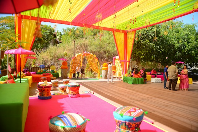 A wedding stage under construction at golden hour, floral artisans arranging marigold and white chrysanthemum panels, ladders and fabric bolts visible
